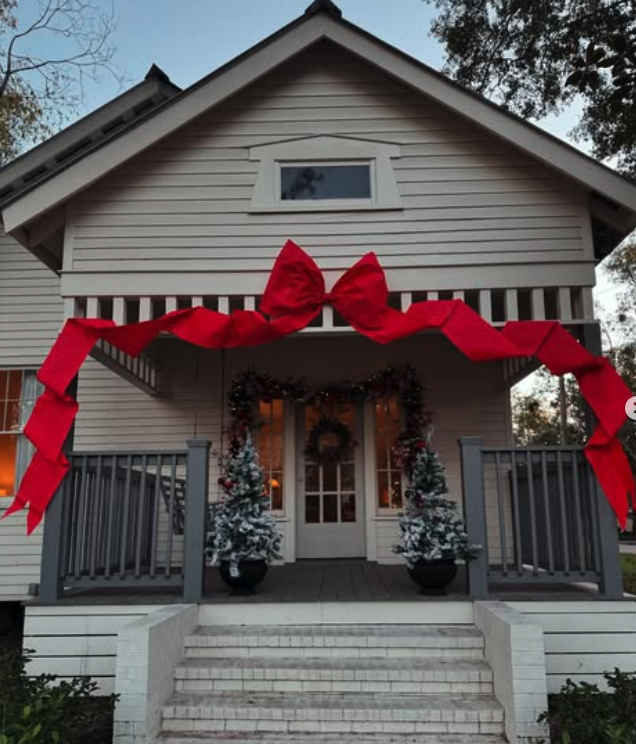 Raised cottage porch with a giant red bow arching over the gable, deep green garland, a wreath, and flocked mini trees in black urns