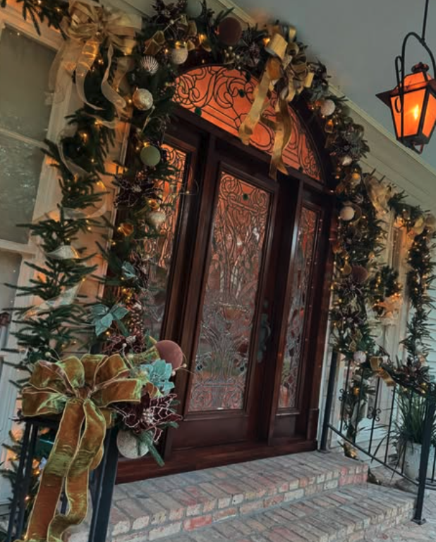 An ironwork front door dressed with garland, wide gold ribbon, bronze and burgundy ornaments, pinecones, and a lit lantern sconce